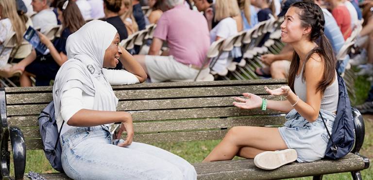 Two students from diverse backgrounds sit on a bench talking